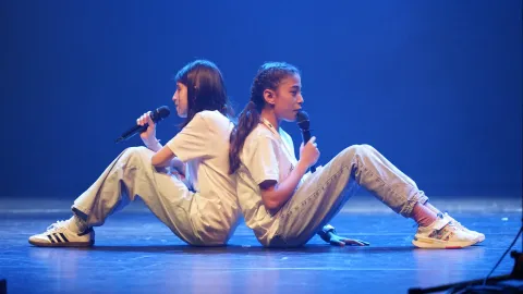 Twee meisjes zitten rug aan rug op het podium en zingen in hun microfoon