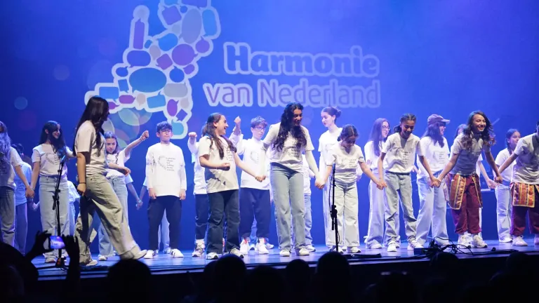 Jongeren staan hand in hand op het podium voor het scherm met het logo van Harmonie van Nederland
