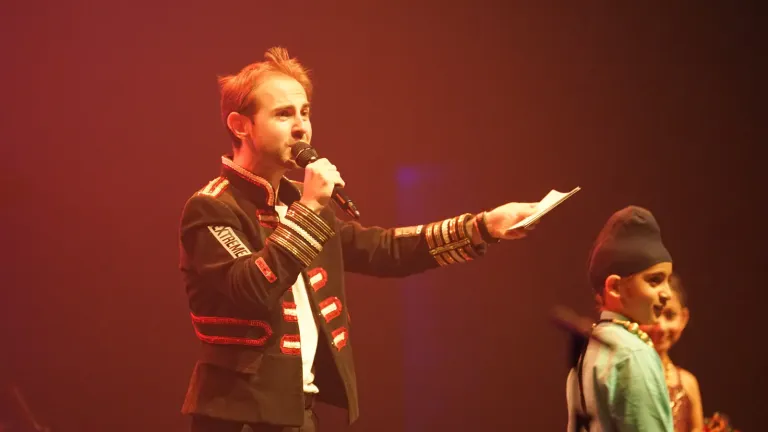 Een man in een zwart jasje met rode bies staat met microfoon op het podium