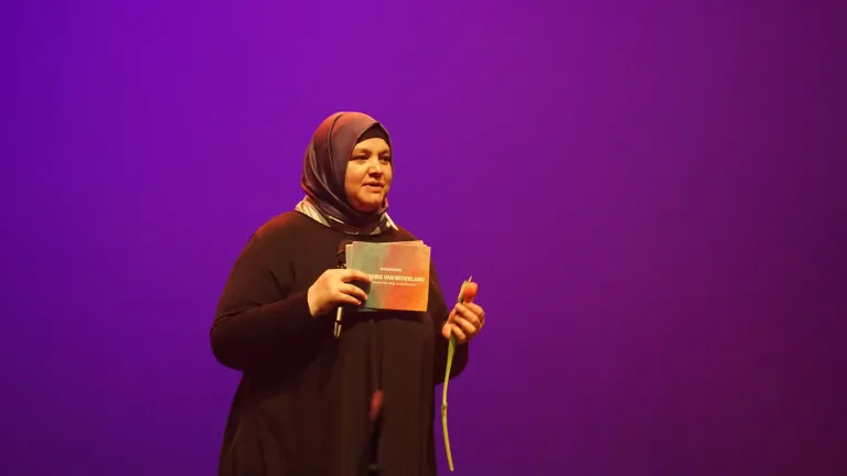 Een vrouw met hoofddoek staat met microfoon en kaartjes op het podium in paars licht