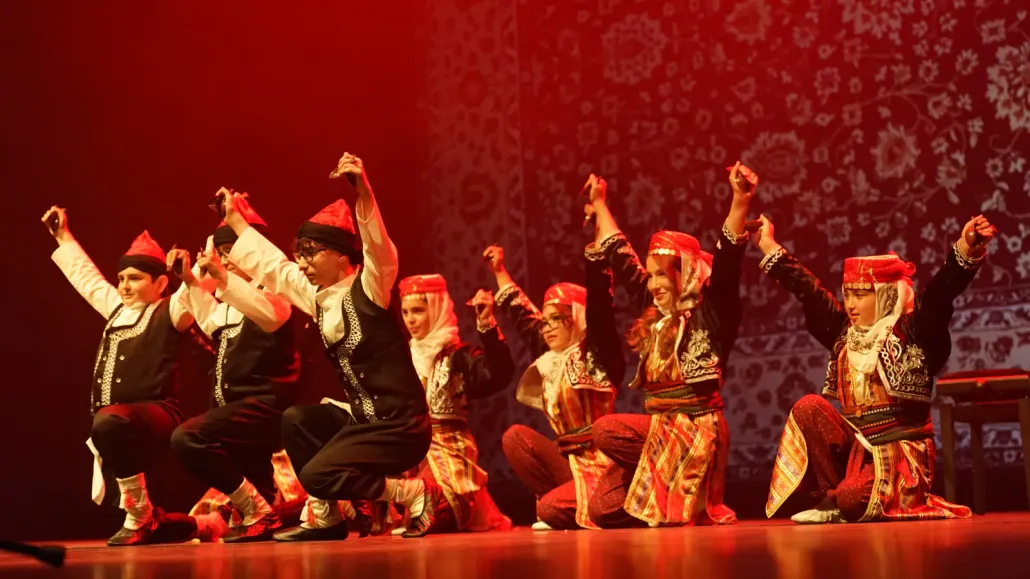 Een dansgroep in traditionele klederdracht met rode hoeden knielt op het podium met opgeheven armen