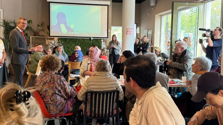 Een spreker praat met een groep bezoekers in een café terwijl een film op het scherm speelt