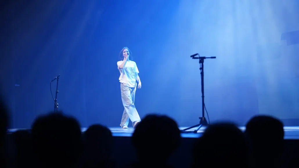 Een meisje zingt op een podium in blauw licht, met het publiek als silhouet op de voorgrond